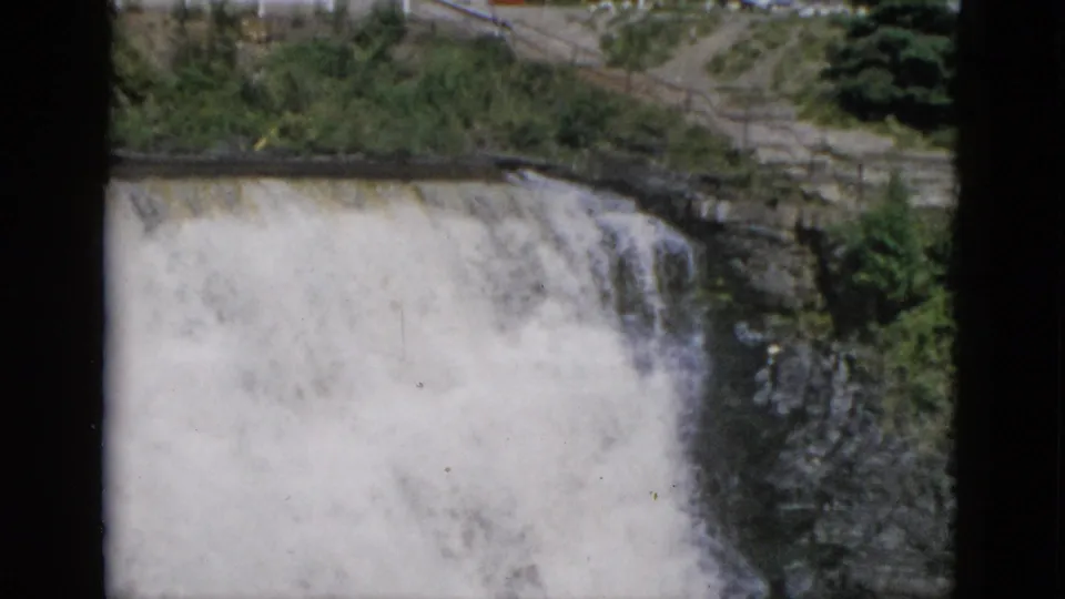 1950s 8mm color film: wide view of a powerful waterfall in Quebec, Canada, with green hillside, dark layered cliff, and roadside infrastructure at the crest.