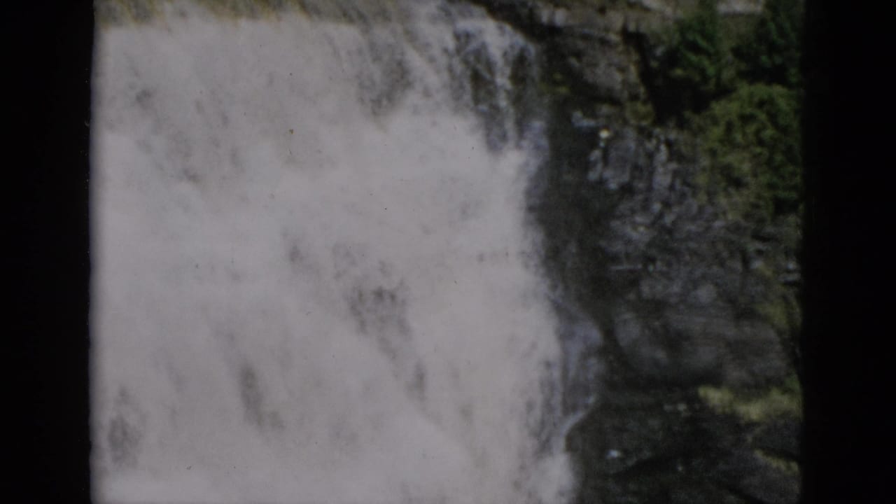 Close view of white water falling against a dark rocky cliff face, green foliage to the right, 8mm film frame borders at edges.