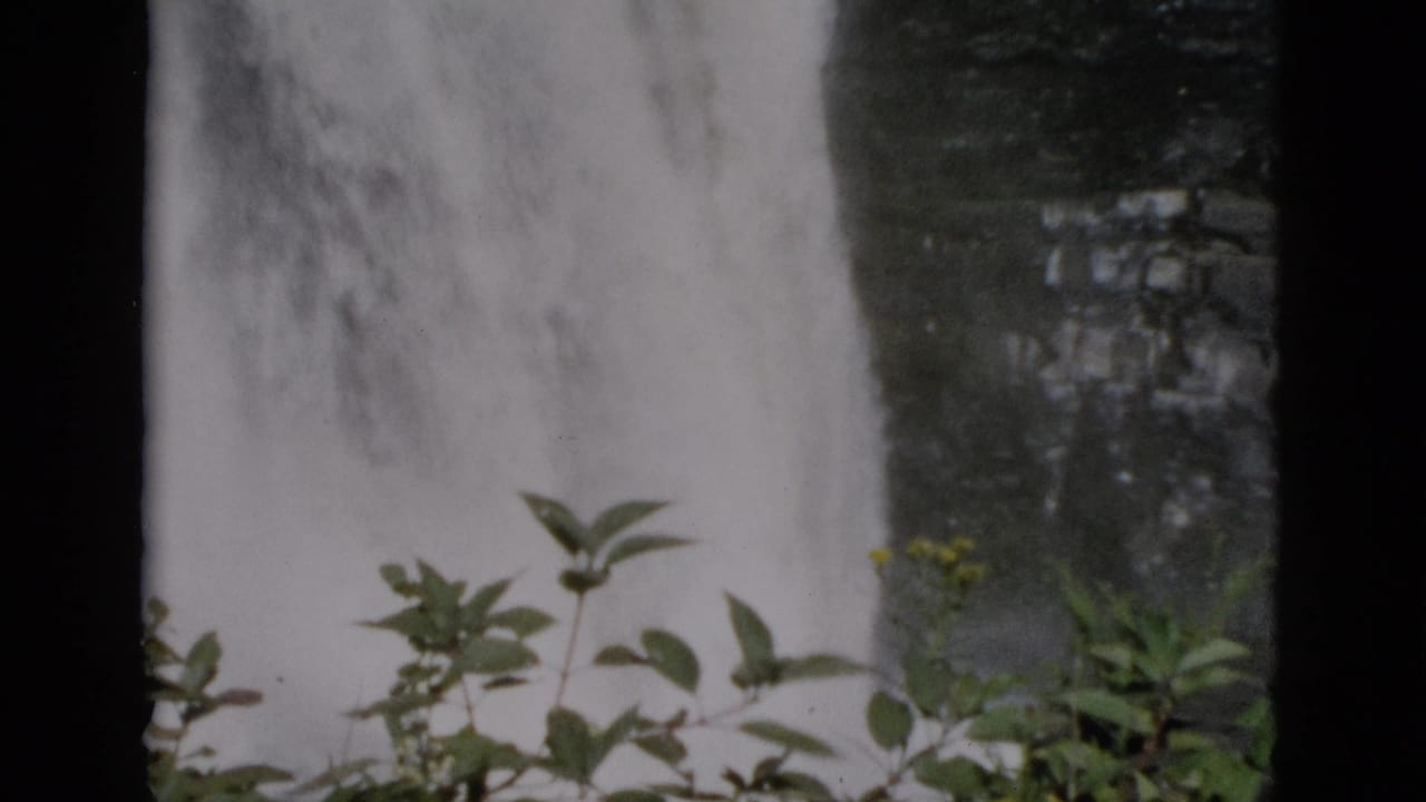 Waterfall over dark rock with green foliage upper right and a flat-roofed building with yellow-framed windows visible at the base.