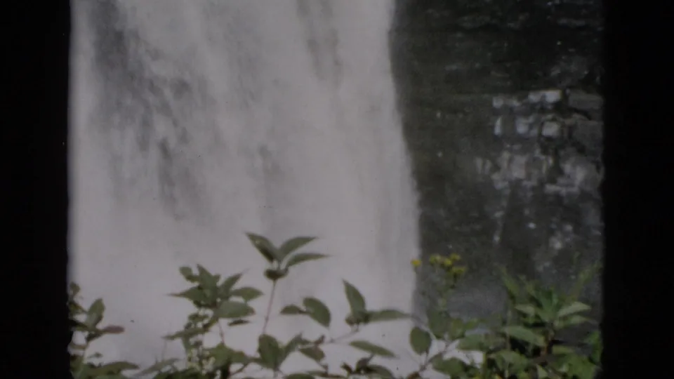 8mm footage of a waterfall alongside a flat-roofed building with yellow-framed windows and green shrubs in the upper right, Quebec, Canada, circa 1952.