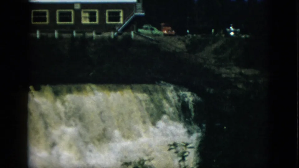 8mm home movie wider shot of a waterfall over a rocky ledge with an adjacent flat-roofed building and a possible vehicle on a hillside, Quebec, Canada, circa 1952.