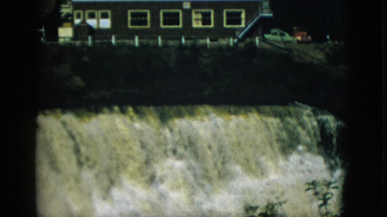 Turbulent waterfall below industrial building; a red or orange period car is visible among parked vehicles on the upper embankment.