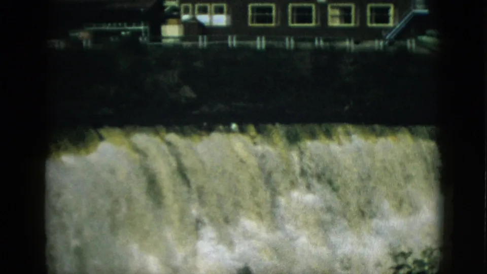 8mm frame circa 1952, Quebec — institutional or industrial building with yellow-framed windows and exterior stairs above a churning waterfall or spillway.