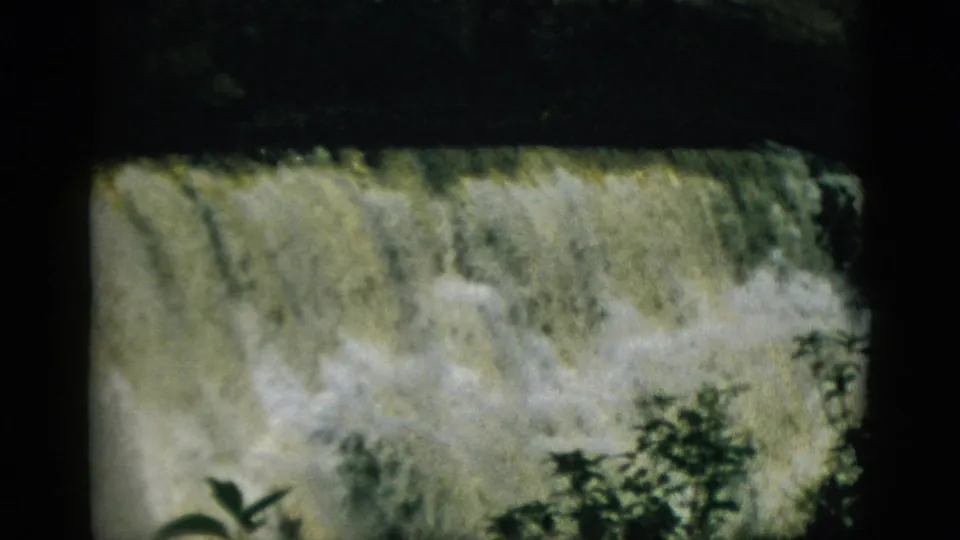 8mm frame circa 1952, Quebec — close view of turbulent waterfall or spillway overflow; heavy white foam and churning water fill the frame with foliage at lower right.