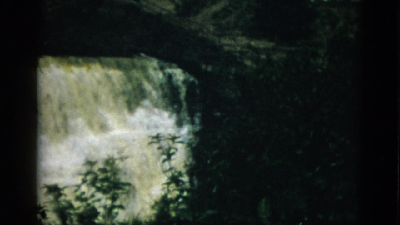 Wide waterfall with white rushing water over a rocky ledge, dark tree silhouettes framing the lower portion, 8mm film.
