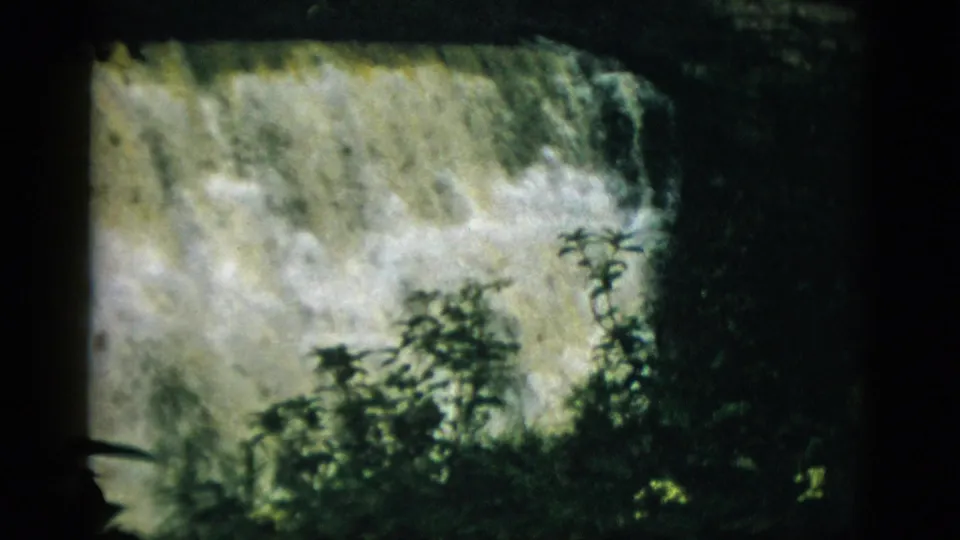 8mm color film, likely 1952 Quebec, with dense dark foreground foliage filling the lower frame and white waterfall cascade visible above and behind.