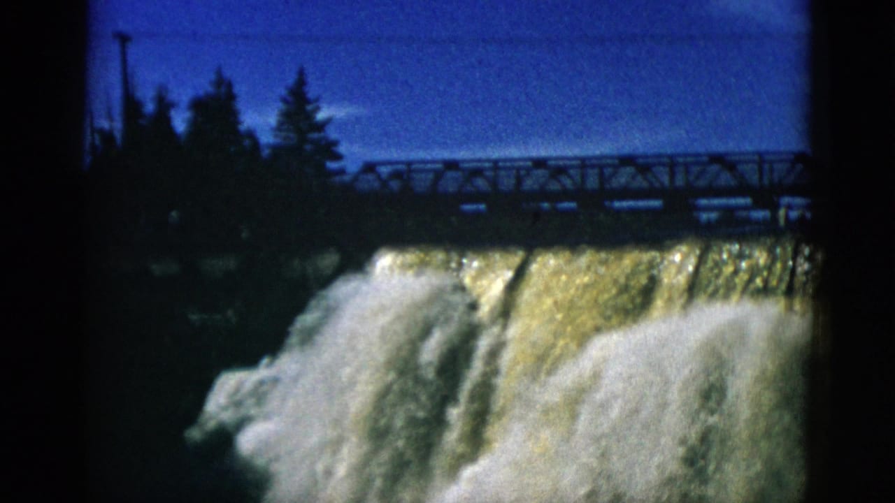 Waterfall with golden-amber water over dam, metal truss bridge at upper right, conifers silhouetted against vivid blue sky.