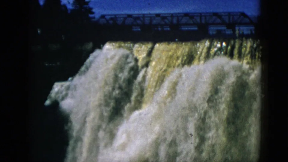 1952 Quebec — 8mm mid-shot of dam waterfall, golden-toned cascading water, white churning base, truss bridge in upper frame against blue sky.