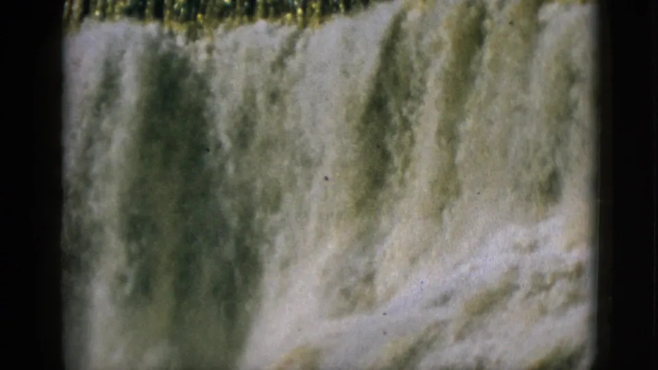 1952 Quebec — close 8mm shot of dam waterfall face, amber and white cascading water, prominent film grain and motion blur, bridge out of frame.
