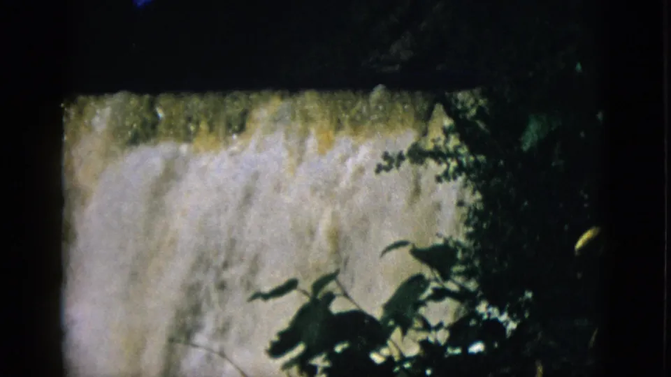 8mm film frame circa 1952 showing a waterfall crest with amber-toned mist and silhouetted foliage at right.