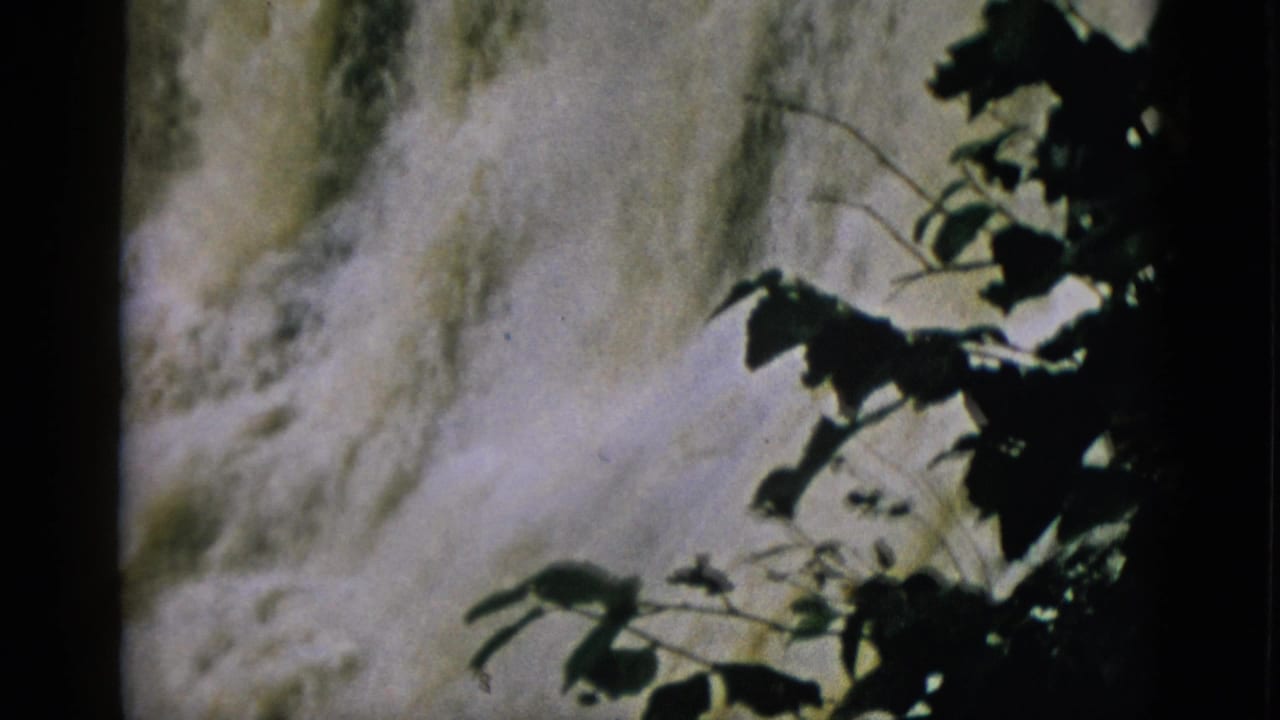 8mm footage from 1952 Quebec — frothy waterfall seen through dark silhouetted tree branches with warm film colour cast.