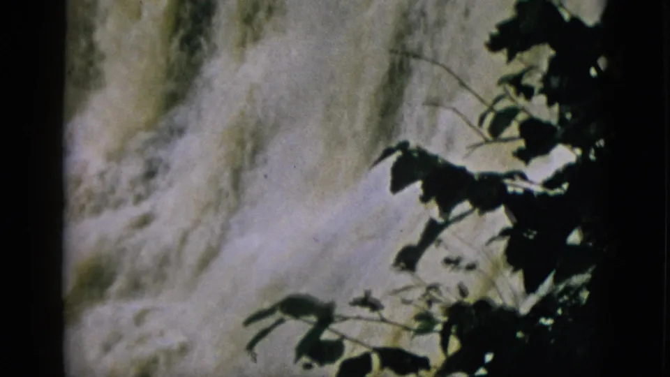 8mm footage from 1952 Quebec — frothy waterfall seen through dark silhouetted tree branches with warm film colour cast.