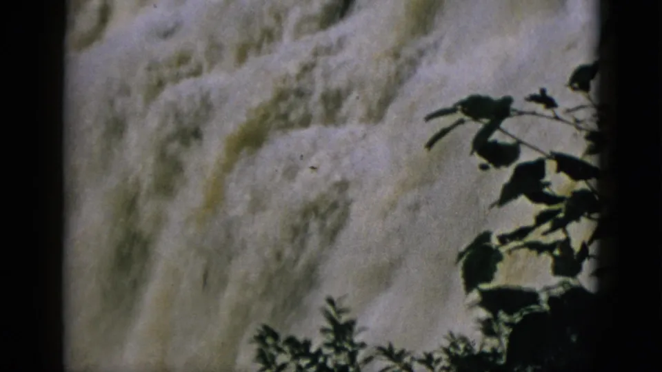 8mm home-movie frame from 1952 Quebec — frothy waterfall behind prominent silhouetted branch and leaf foreground.