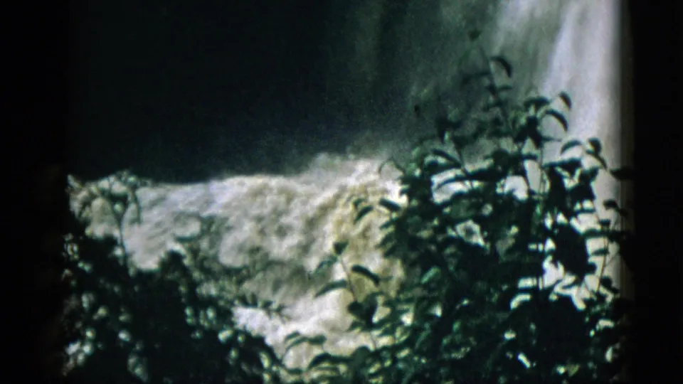 Pulled-back 8mm view from 1952 Quebec showing waterfall through green shrubs with dark overcast sky or mist visible above.