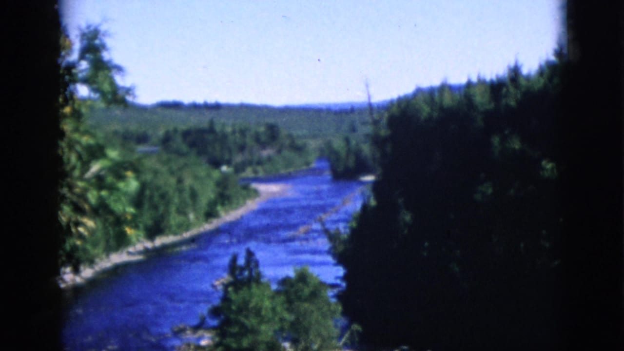 Wide blue river between dense forest banks, pan continues, pale blue sky, film-gate border at left edge.