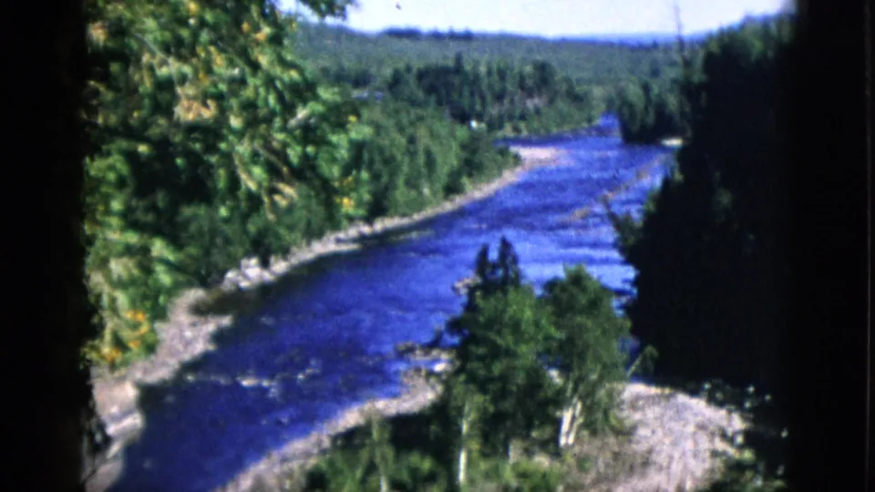 Near-bank gravel and light-trunked trees framing a blue river; dense boreal forest on far bank; Quebec, Canada, circa 1952, 8mm home movie.