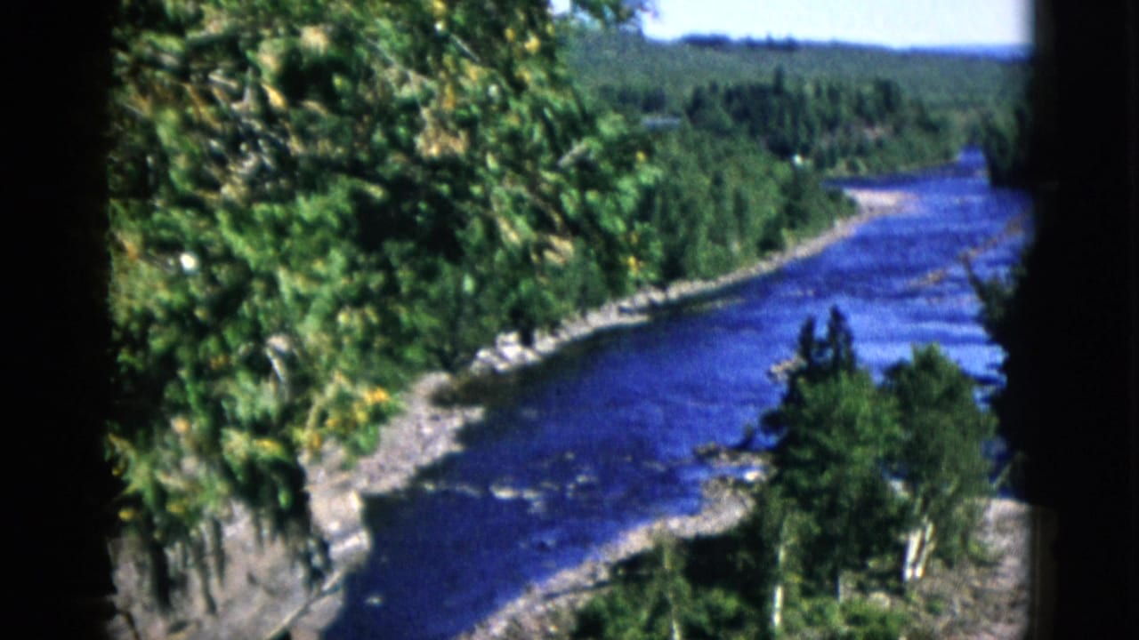 Blue river winding through dense spruce forest with rocky shoreline, viewed from elevated position on 8mm film.