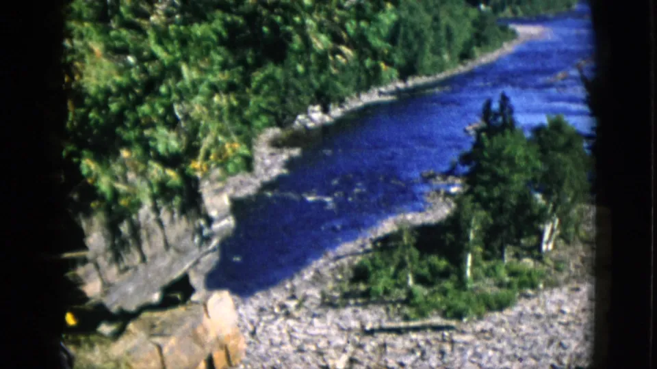 8mm film c.1952, Quebec — pan reveals fallen timber in foreground as river curves through dense coniferous forest with gravel shore.