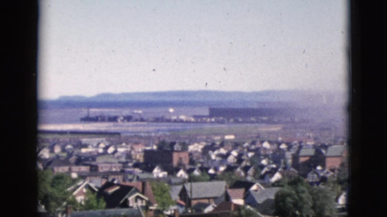 8mm color film, Quebec, Canada, 1952: densely packed residential rooftops with scattered deciduous trees and a wide flat expanse visible in the distant background.
