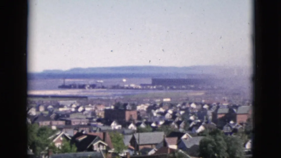 8mm color film, Quebec, Canada, 1952: densely packed residential rooftops with scattered deciduous trees and a wide flat expanse visible in the distant background.