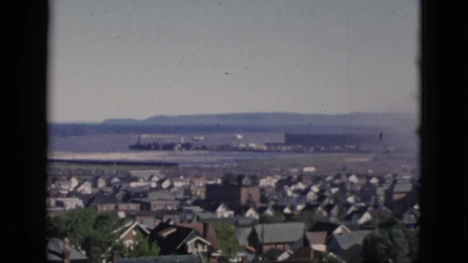 8mm color film frame from Quebec, Canada, circa 1952: panoramic hilltop view over a mid-century residential area with a wide flat expanse and low hills in the distance.