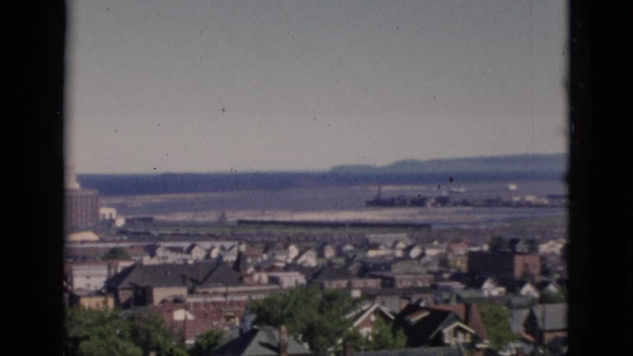 8mm home-movie pan start over a dense residential neighbourhood, likely Quebec, Canada, circa 1952, with foreground deciduous trees, varied rooftops, and a distant river.