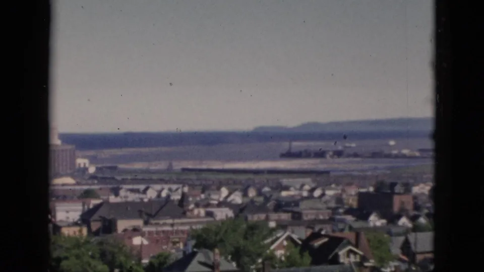 8mm home-movie pan start over a dense residential neighbourhood, likely Quebec, Canada, circa 1952, with foreground deciduous trees, varied rooftops, and a distant river.