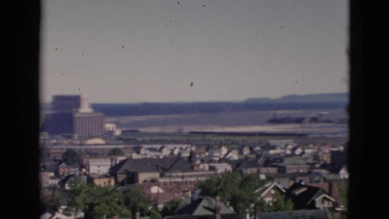 Pan moved left to reveal a large multi-story institutional building near the waterfront, beyond rows of residential rooftops and foreground trees.