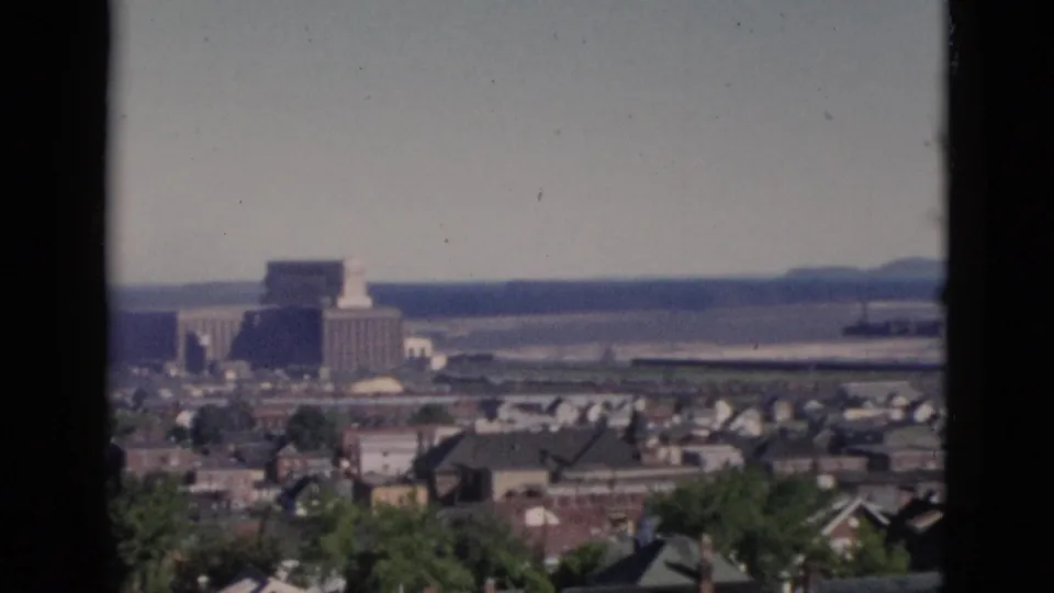 Continued 8mm pan over Quebec residential district circa 1952, with a prominent multi-storey institutional building now centred against the river and low rolling hills.