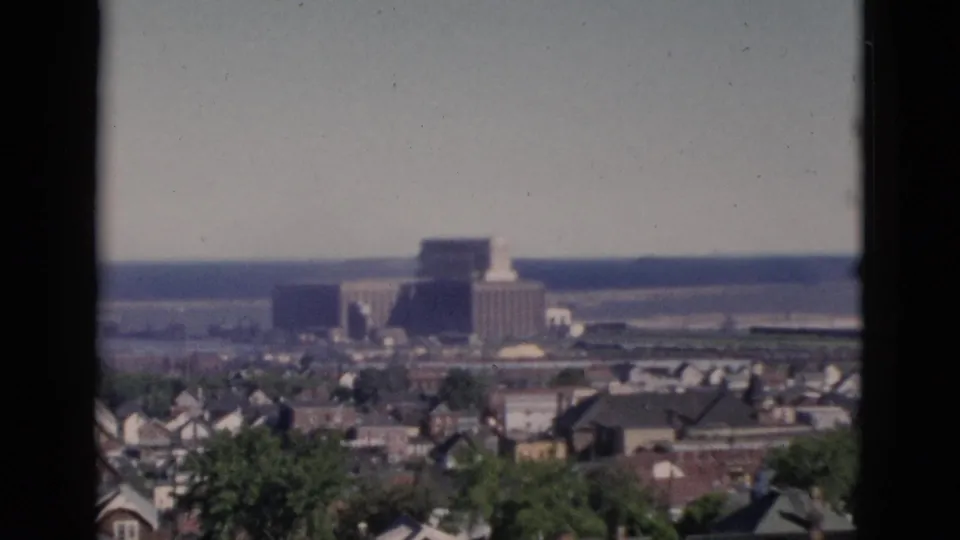 Elevated view of a dense residential neighborhood in Quebec, Canada, circa 1952, with pitched-roof houses, green trees, and a large building on the horizon, on 8mm color film.