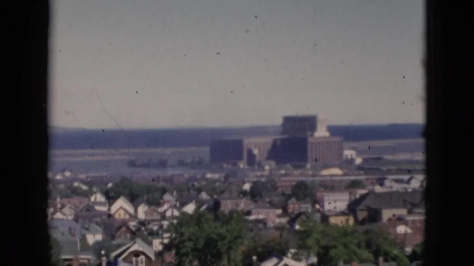 Elevated cityscape view consistent with Quebec, Canada, circa 1952, showing residential rooftops, green trees, and a large building in the mid-distance under a hazy sky, on 8mm color film.
