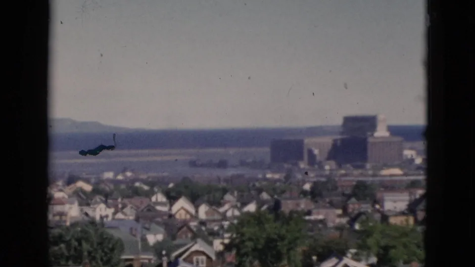 Elevated panorama likely in Quebec, Canada, circa 1952, with residential rooftops spreading to the horizon and a large block building in the mid-distance under a hazy sky, on 8mm color film.