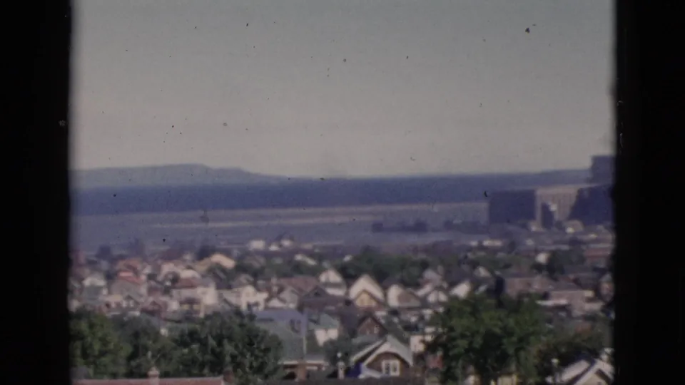 Elevated cityscape view consistent with Quebec, Canada, circa 1952, showing residential rooftops in the foreground and a large building with a distant landscape feature on the right horizon, on 8mm color film.