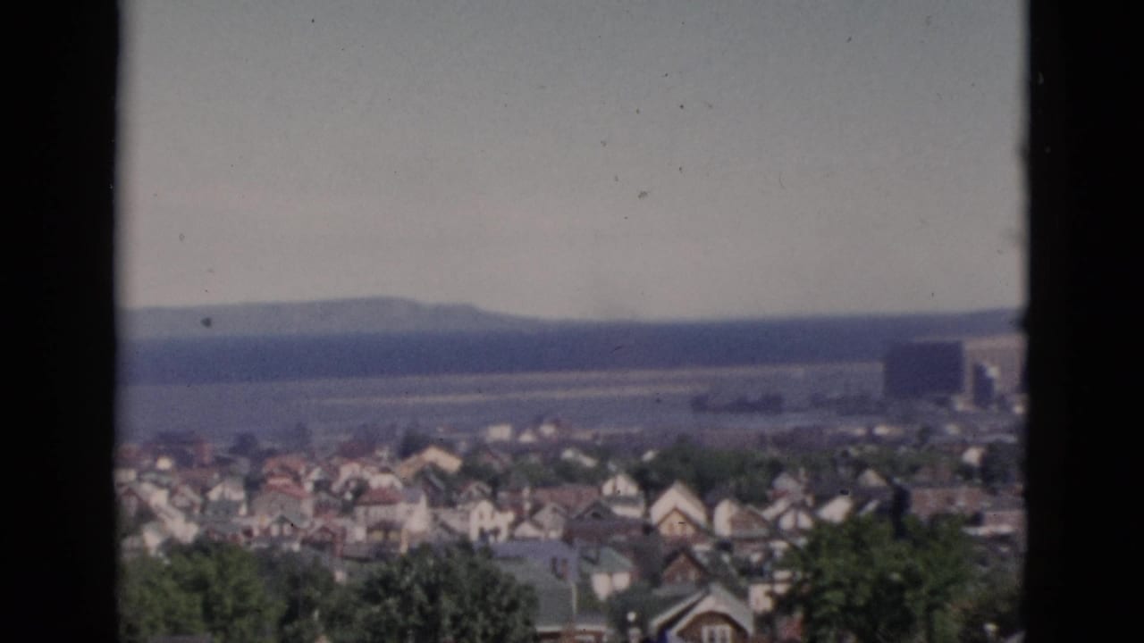 Window-framed panorama of a Quebec coastal town circa 1952, showing tree-lined residential streets and a wide body of water on the horizon