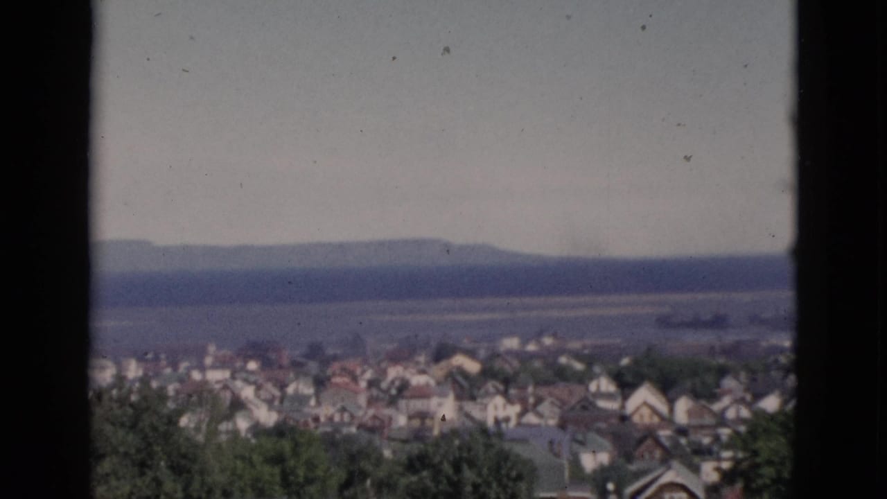Static wide shot of Quebec townscape circa 1952, window-framed residential rooftops and tree canopy with a distant body of water visible on the horizon