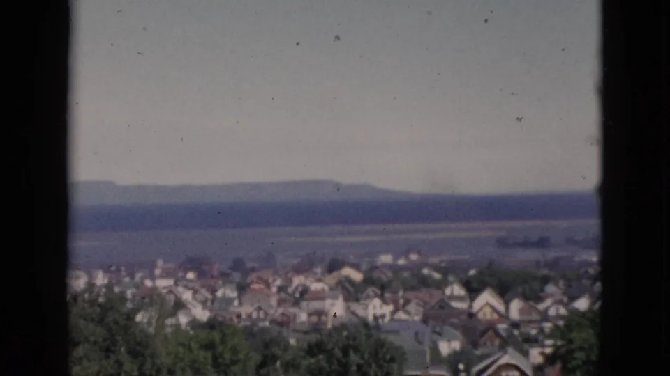 Static wide shot of Quebec townscape circa 1952, window-framed residential rooftops and tree canopy with a distant body of water visible on the horizon