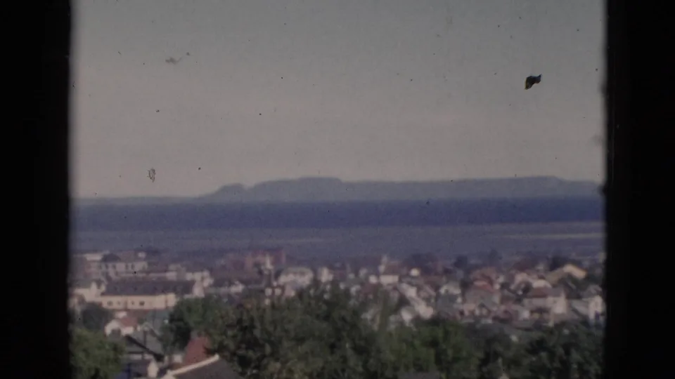 Window-framed view of Quebec coastal town circa 1952, wide residential rooftop panorama with distant waterway on the horizon and the irregular white form no longer present