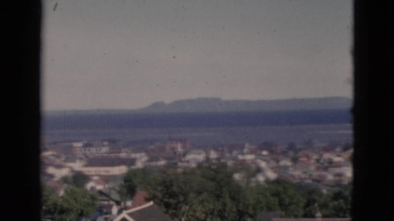 Elevated view of a Quebec coastal community circa 1952, showing residential rooftops, green trees, dense townscape, and a wide water body with distant hills, shot on 8mm film.