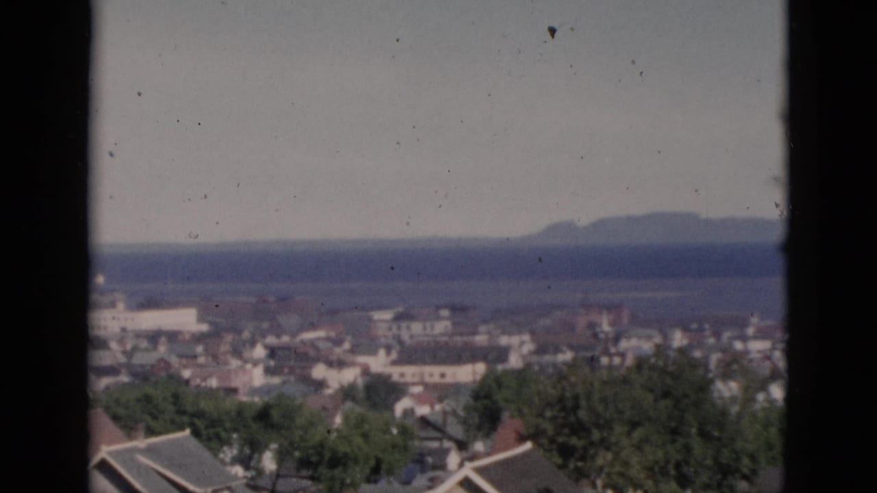 Elevated 8mm film view of a mid-century coastal town showing rooftops, trees, townscape, and a wide water body.