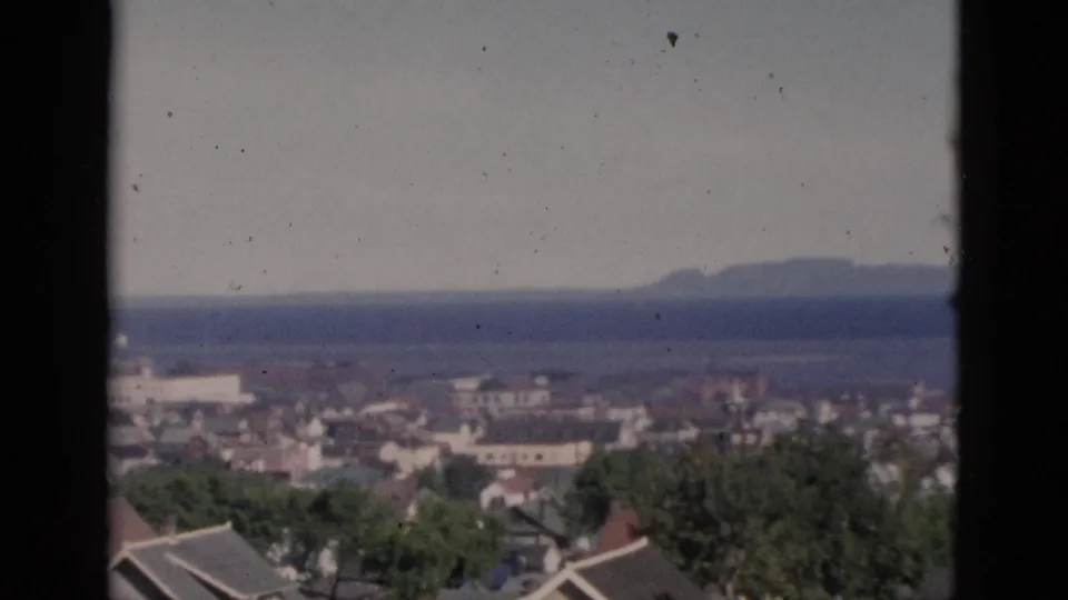 Elevated view of a Quebec coastal community circa 1952; rooftops and trees in foreground, wide water body and distant headland beyond, on 8mm colour film with natural grain and colour fading.