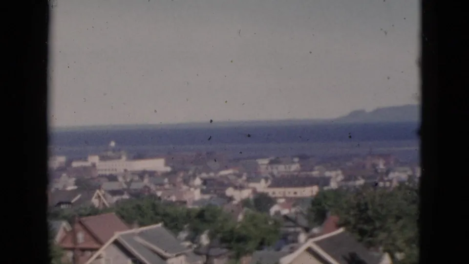 Elevated view circa 1952 of a Quebec coastal town; more rooftop detail visible as the framing shifts slightly downward, water body and distant hills remain in background on 8mm colour film.