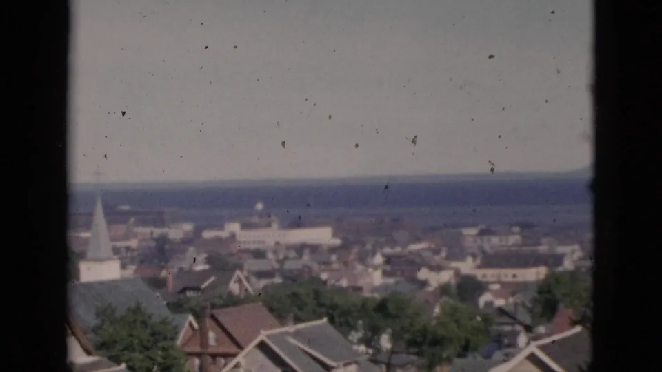 Elevated view circa 1952 of a Quebec coastal town; a church steeple rises among residential rooftops in the mid-ground, with a wide water body and distant headland beyond, on 8mm colour film.