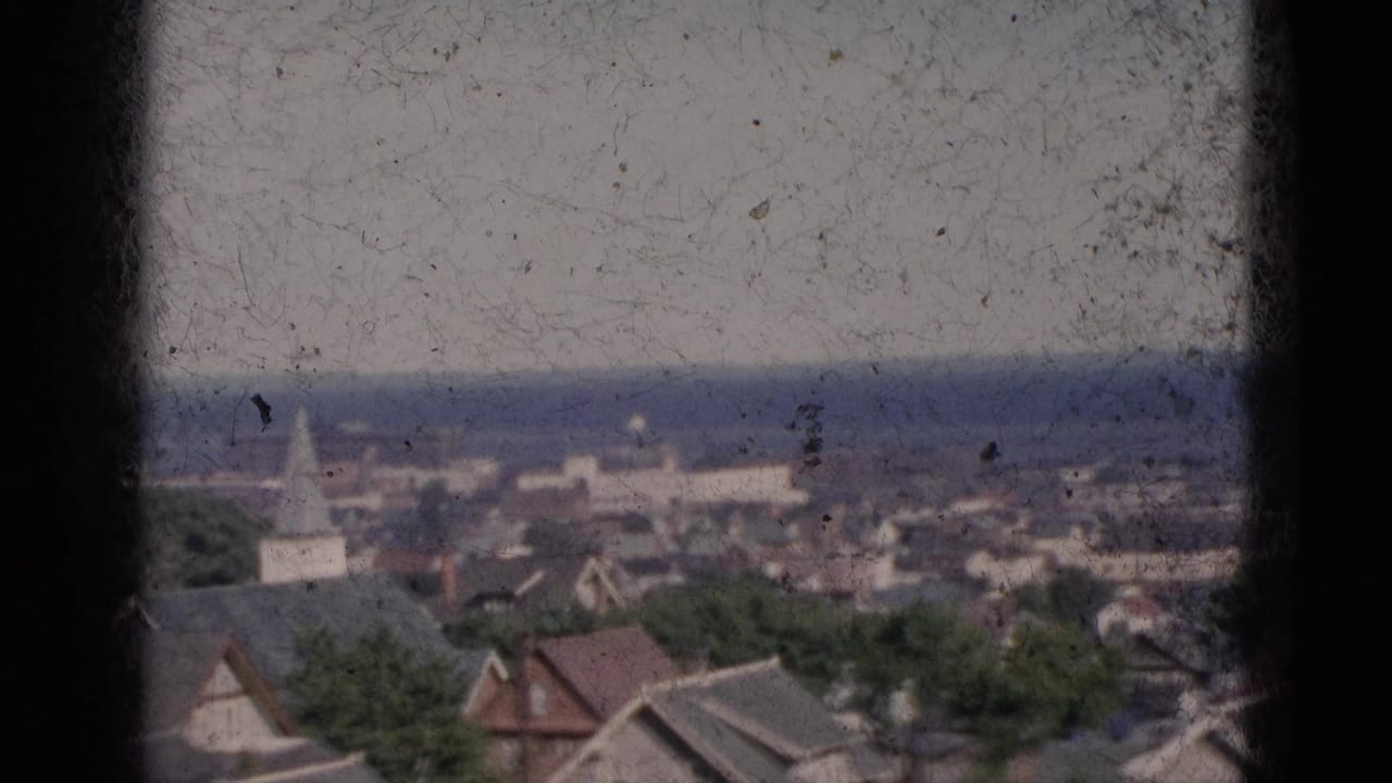 Circa-1952 8mm panorama of a Quebec town viewed through a window frame, showing residential rooftops, a white church steeple, green trees, and a distant body of water.