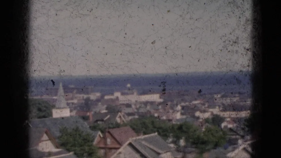 Circa-1952 8mm panorama of a Quebec town viewed through a window frame, showing residential rooftops, a white church steeple, green trees, and a distant body of water.