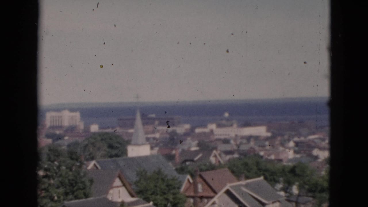 Circa-1952 8mm color footage of a Quebec town from an elevated window, with residential rooftops, a white steeple, green trees, a rectangular background structure, and a distant waterway.