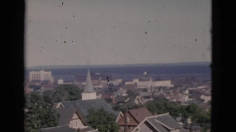 Circa-1952 8mm color footage of a Quebec town from an elevated window, with residential rooftops, a white steeple, green trees, a rectangular background structure, and a distant waterway.