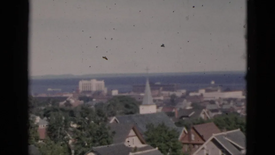 Circa-1952 8mm frame showing Quebec rooftops closer to camera, with a white church steeple, green summer trees, a large background structure, and a pale body of water on the horizon.