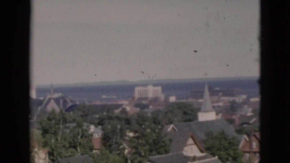 Circa-1952 8mm color footage over Quebec town rooftops, with a white steeple, green trees, and a body of water stretching to the horizon under a pale sky.