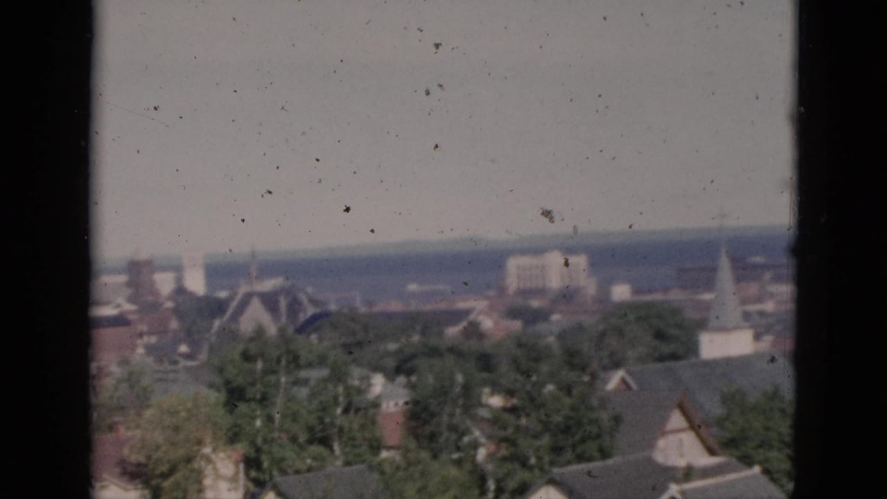 Elevated panorama of what may be a Quebec town, circa 1952, showing a church steeple, brick rooftops, and mature trees on color 8mm film with dust and grain.