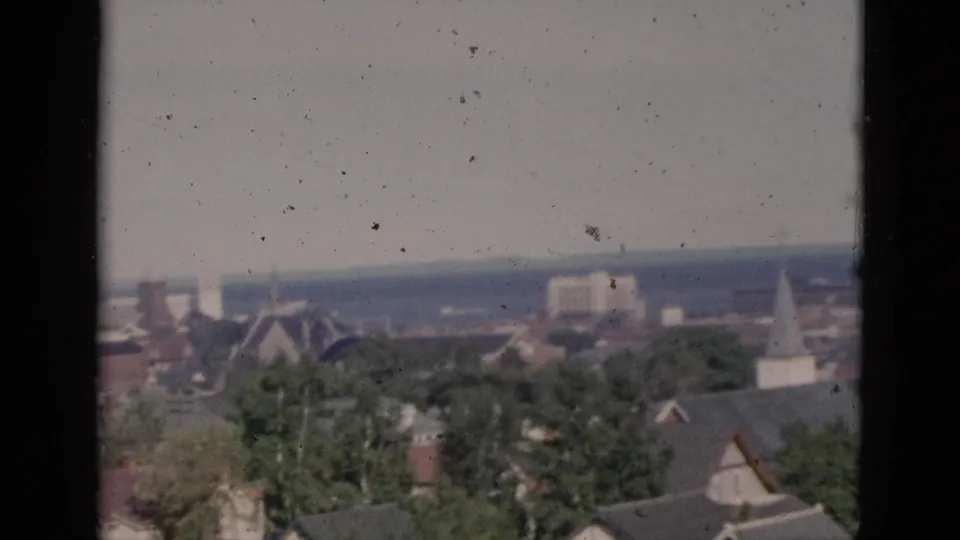Elevated panorama of what may be a Quebec town, circa 1952, showing a church steeple, brick rooftops, and mature trees on color 8mm film with dust and grain.